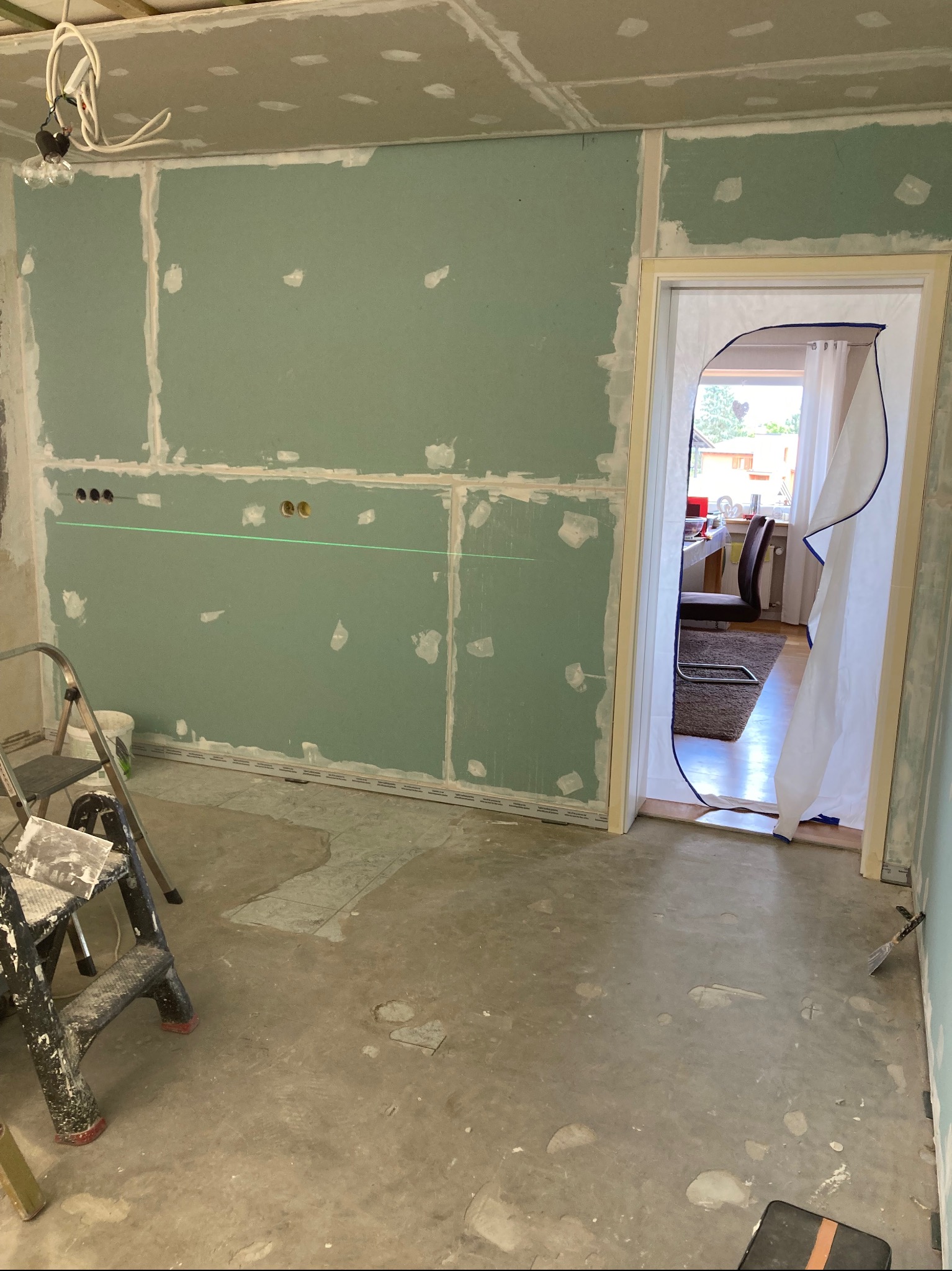 Wnętrze w trakcie prac: ściany z płyty karton-gips, widoczny laser poziomujący, drabina malarska. Otwór drzwiowy zabezpieczony folią. Podłoga surowa, przygotowana do dalszych prac.