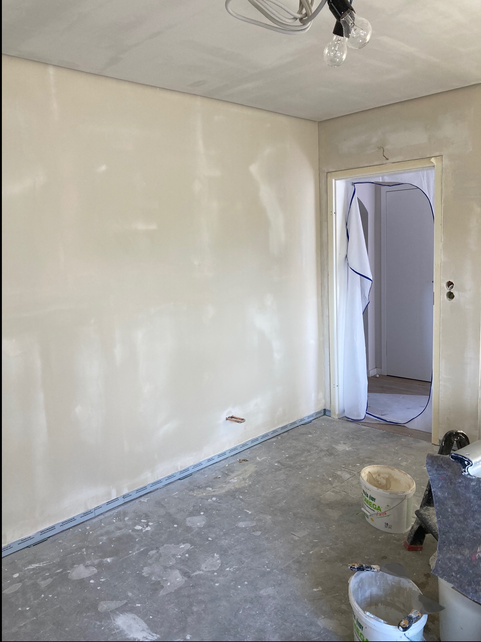 Pokój po szpachlowaniu ścian, widoczne narzędzia, wiaderka z masą i zabezpieczona folią futryna drzwiowa. Ściany przygotowane do dalszych prac wykończeniowych.