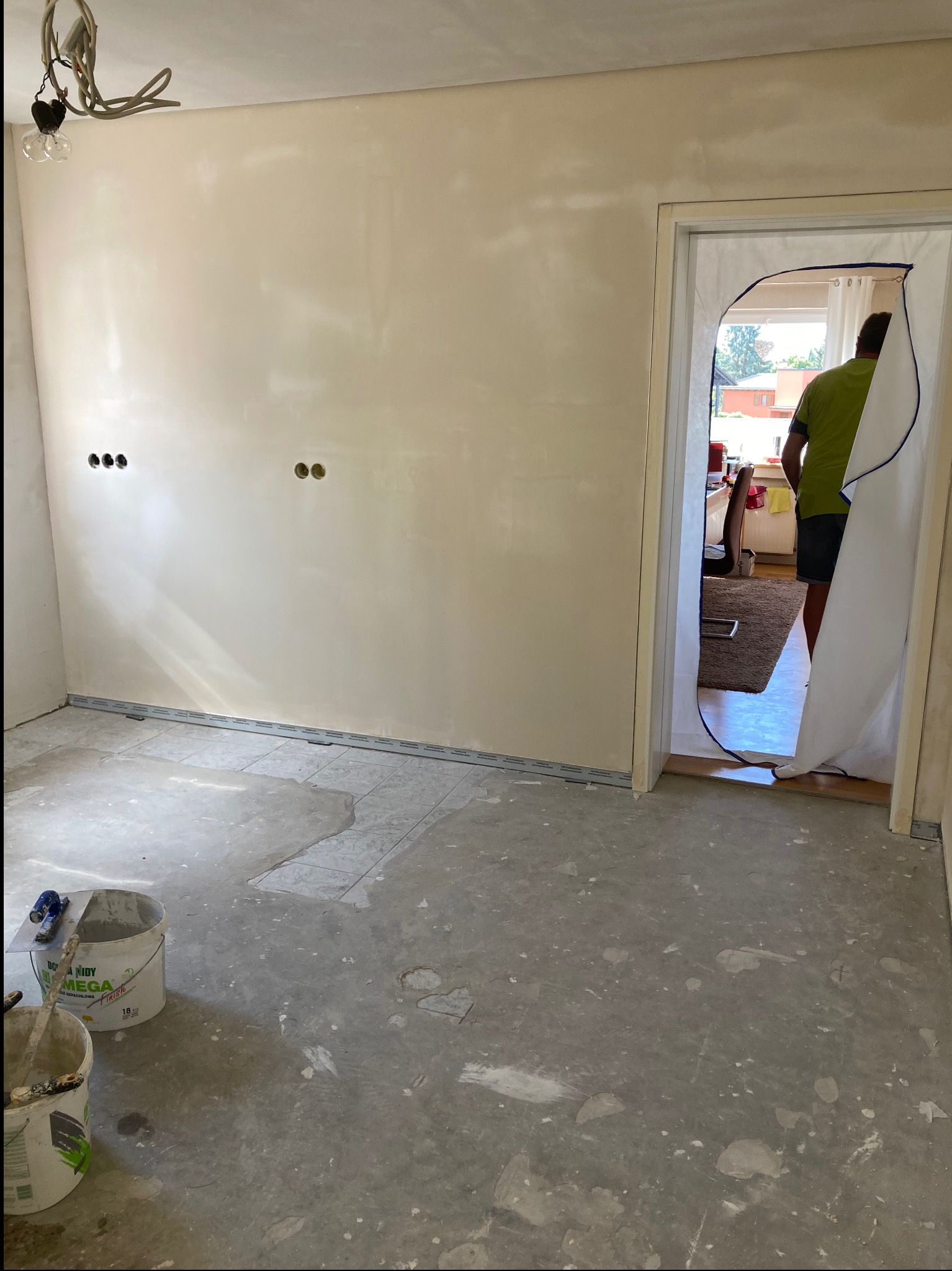 Pokój w trakcie prac wykończeniowych: ściany po szpachlowaniu, widoczne puszki elektryczne, zakryte drzwi folią ochronną, wiaderka z zaprawą na podłodze.