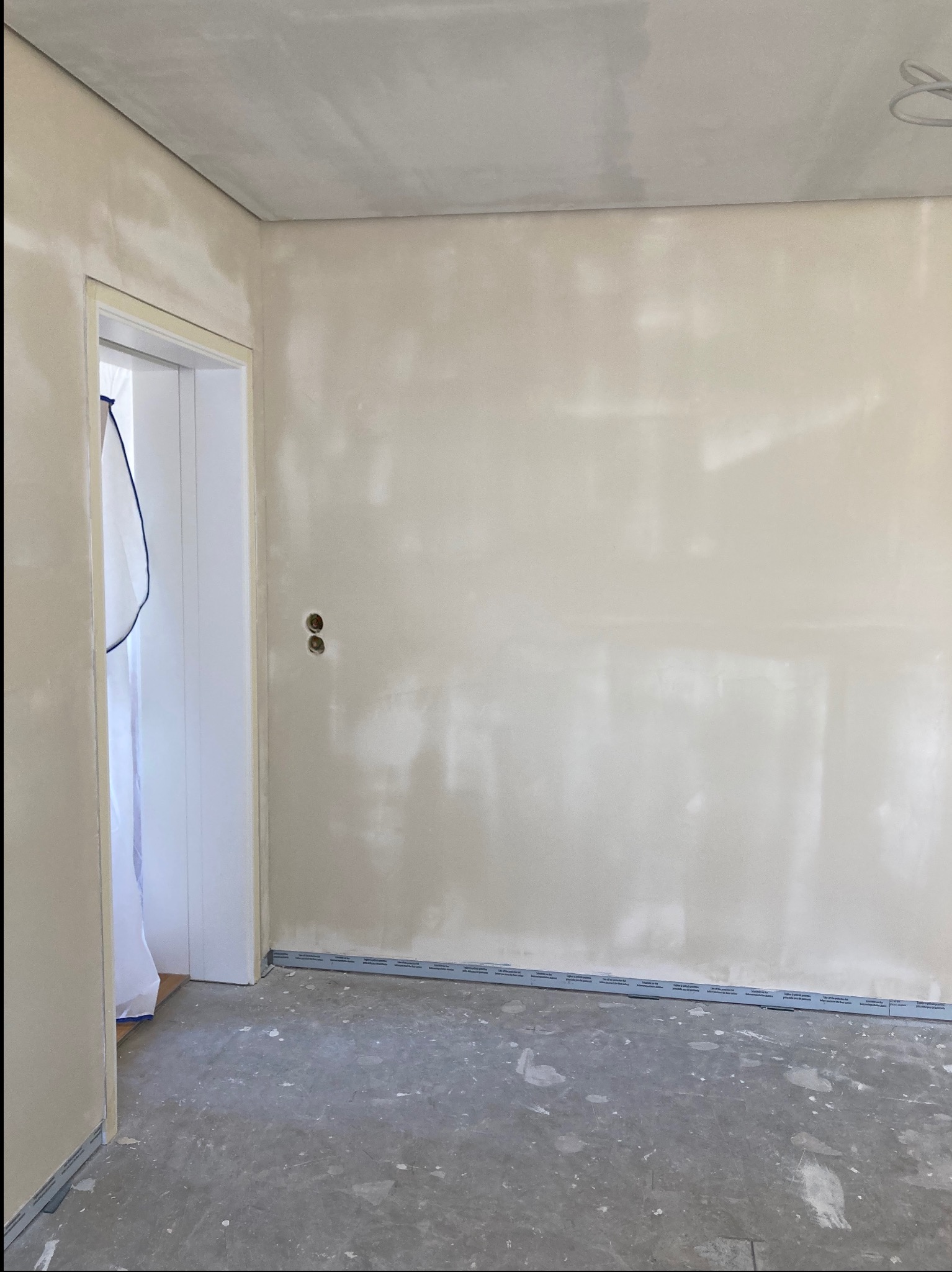 Pokój po szpachlowaniu ścian i sufitu na gładko, widoczne profile przypodłogowe, zabezpieczone drzwi folią. Surowy beton na podłodze.