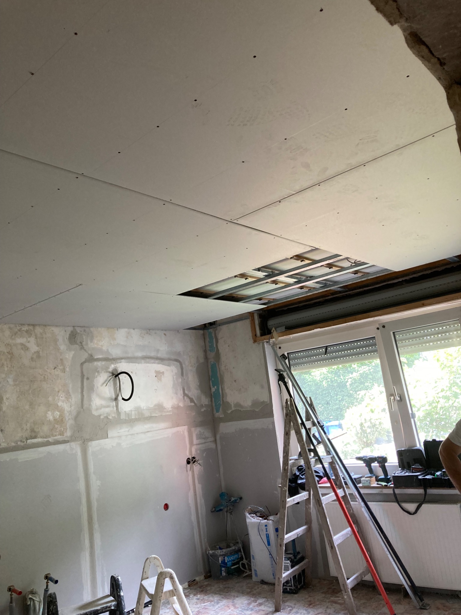 Montaż sufitu podwieszanego z płyt kartonowo-gipsowych w pomieszczeniu z oknem. Widoczne drabiny i narzędzia. Częściowo wykończona ściana z instalacją elektryczną.