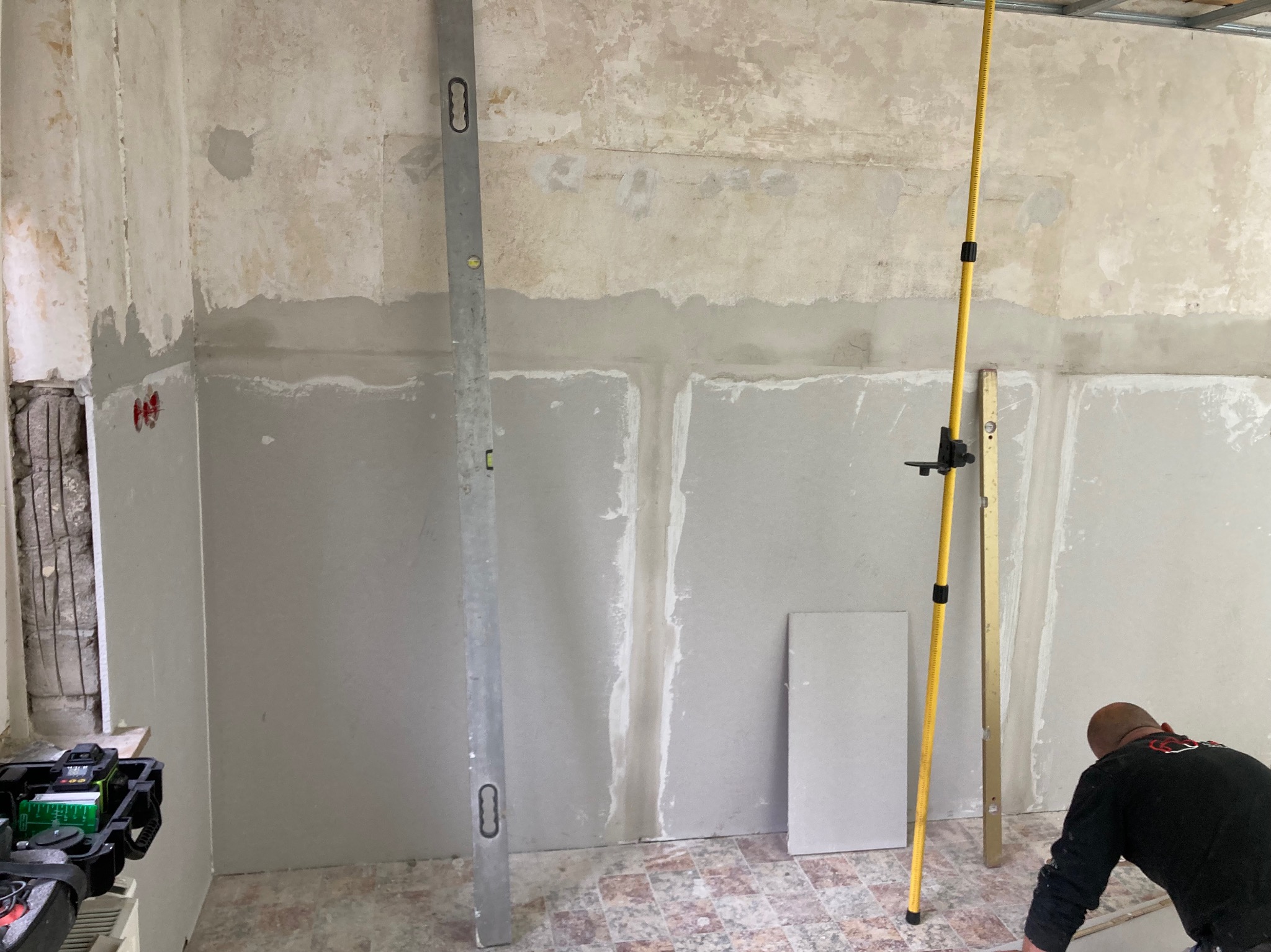 Przygotowanie ścian pod wykończenie: częściowo nałożona gładź, poziomice, laser krzyżowy. Widoczne płyty kartonowo-gipsowe i fragment instalacji elektrycznej.