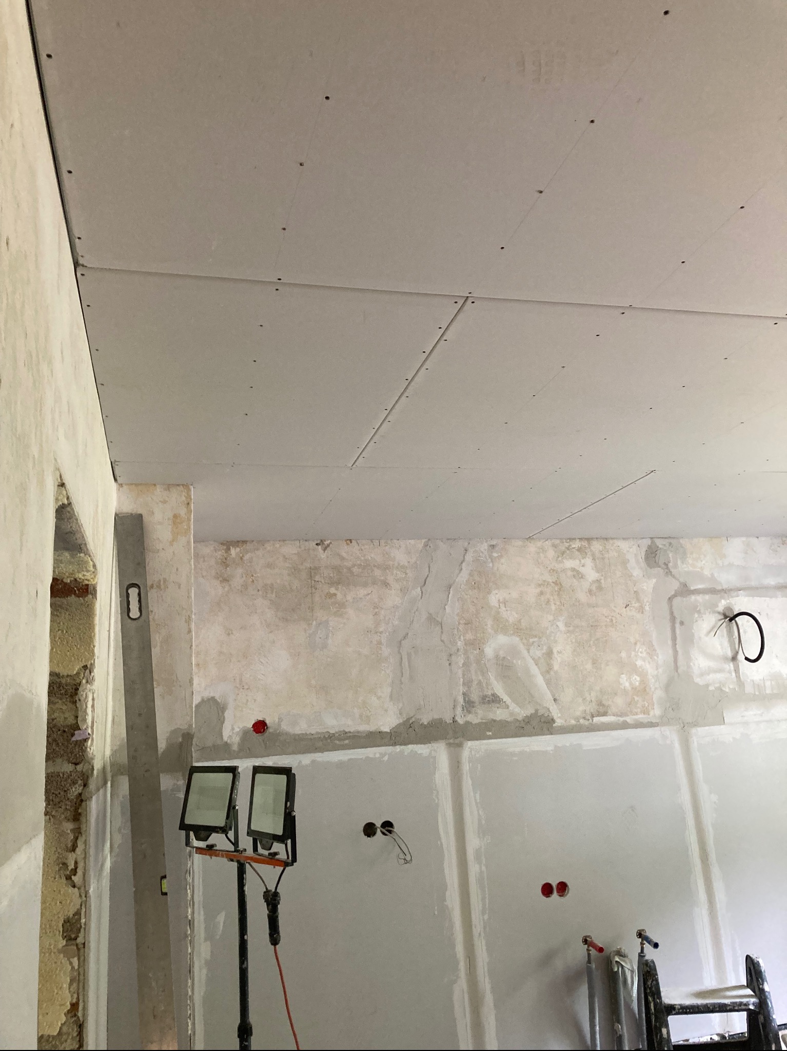 Montaż płyt karton-gips na suficie, widoczne łączenia i wkręty. Ściany w trakcie przygotowania, widoczne ślady po instalacjach. Na podłodze drabina i lampy budowlane.
