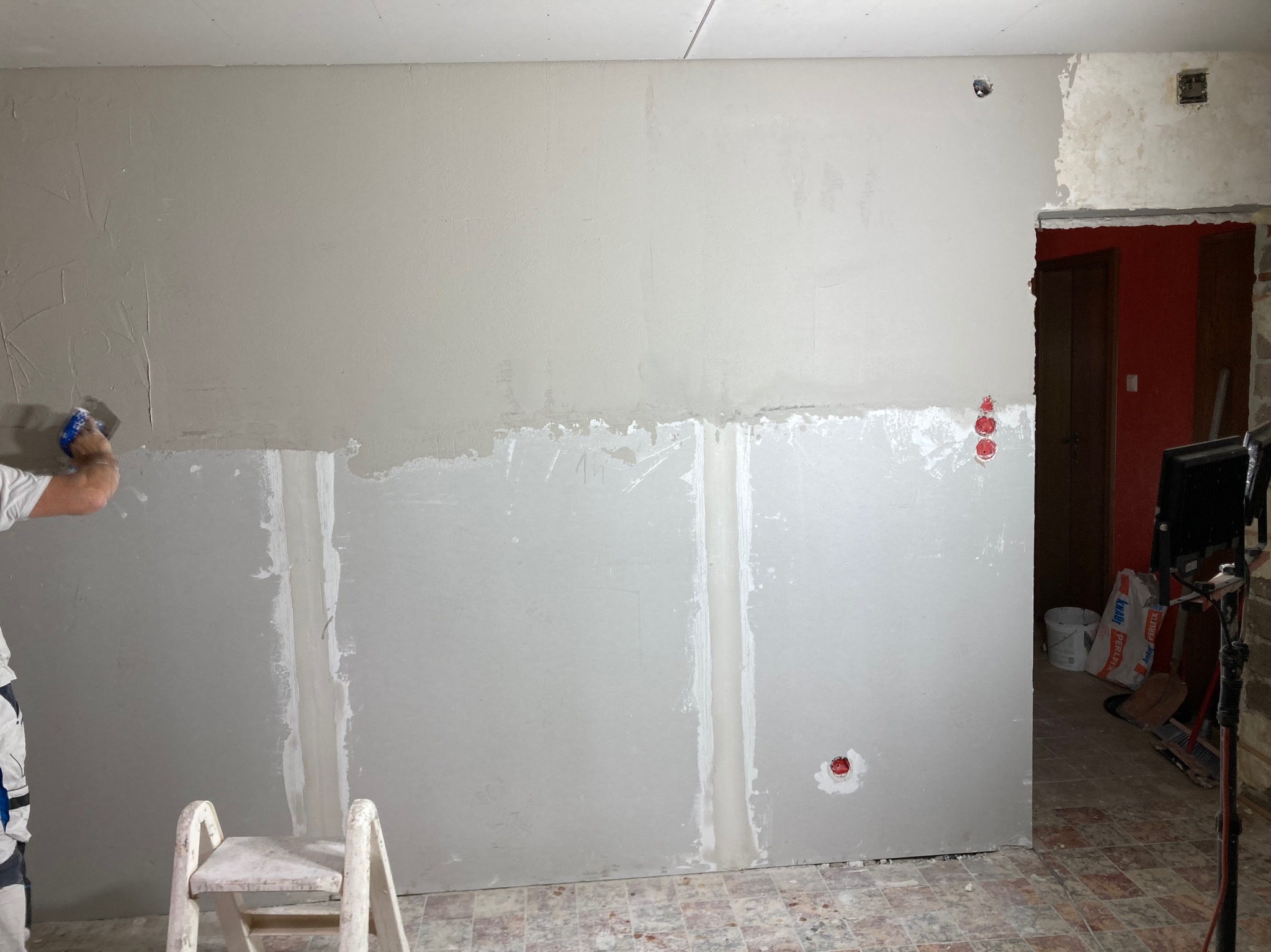 Ściana w trakcie szpachlowania, widoczne łączenia płyt karton-gips, pracownik w kombinezonie nanosi gładź szpachlą. Fragment pokoju z widocznym wejściem do innego pomieszczenia.