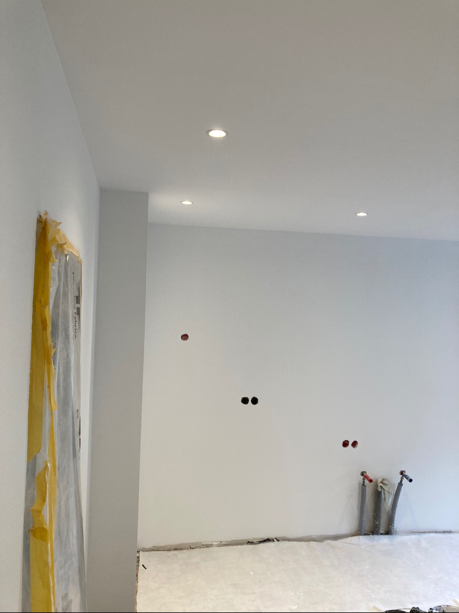 Wnętrze po remoncie: białe ściany z widocznymi punktami elektrycznymi i rurami, zabezpieczone drzwi folią. Minimalistyczne oświetlenie sufitowe.