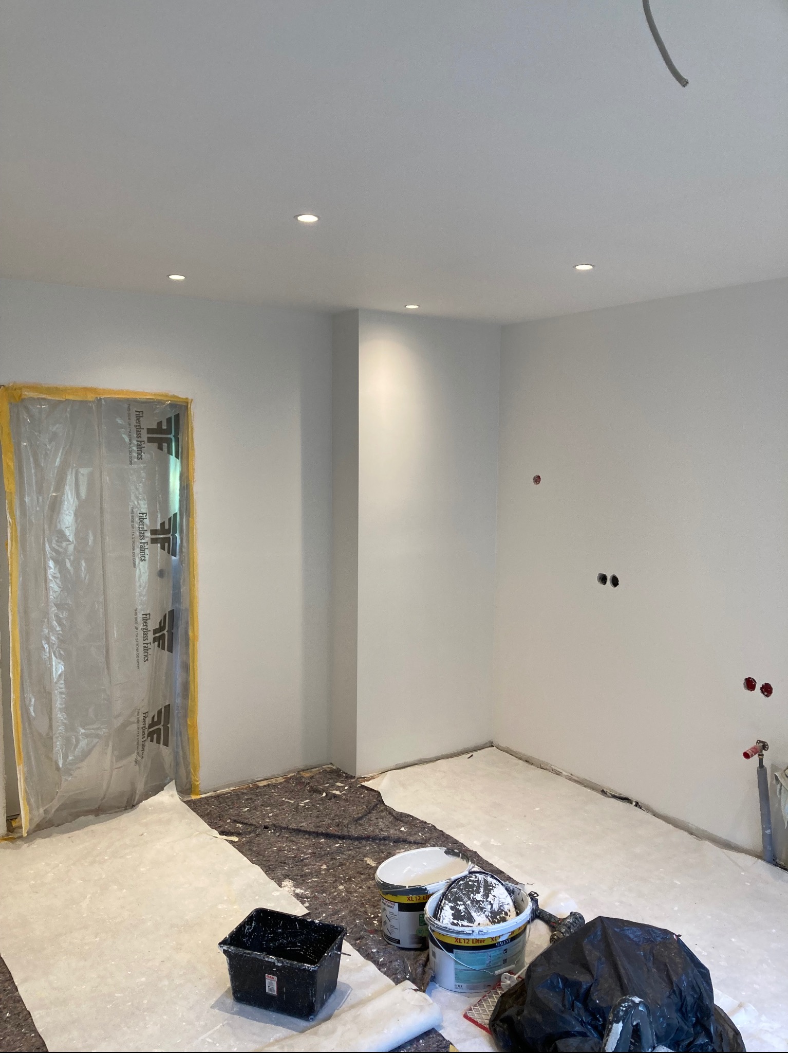 Pokój po szpachlowaniu, przygotowany do malowania: zabezpieczone drzwi folią, podłoga przykryta, widoczne puszki z farbą, ściany gładkie, gotowe do nałożenia koloru.