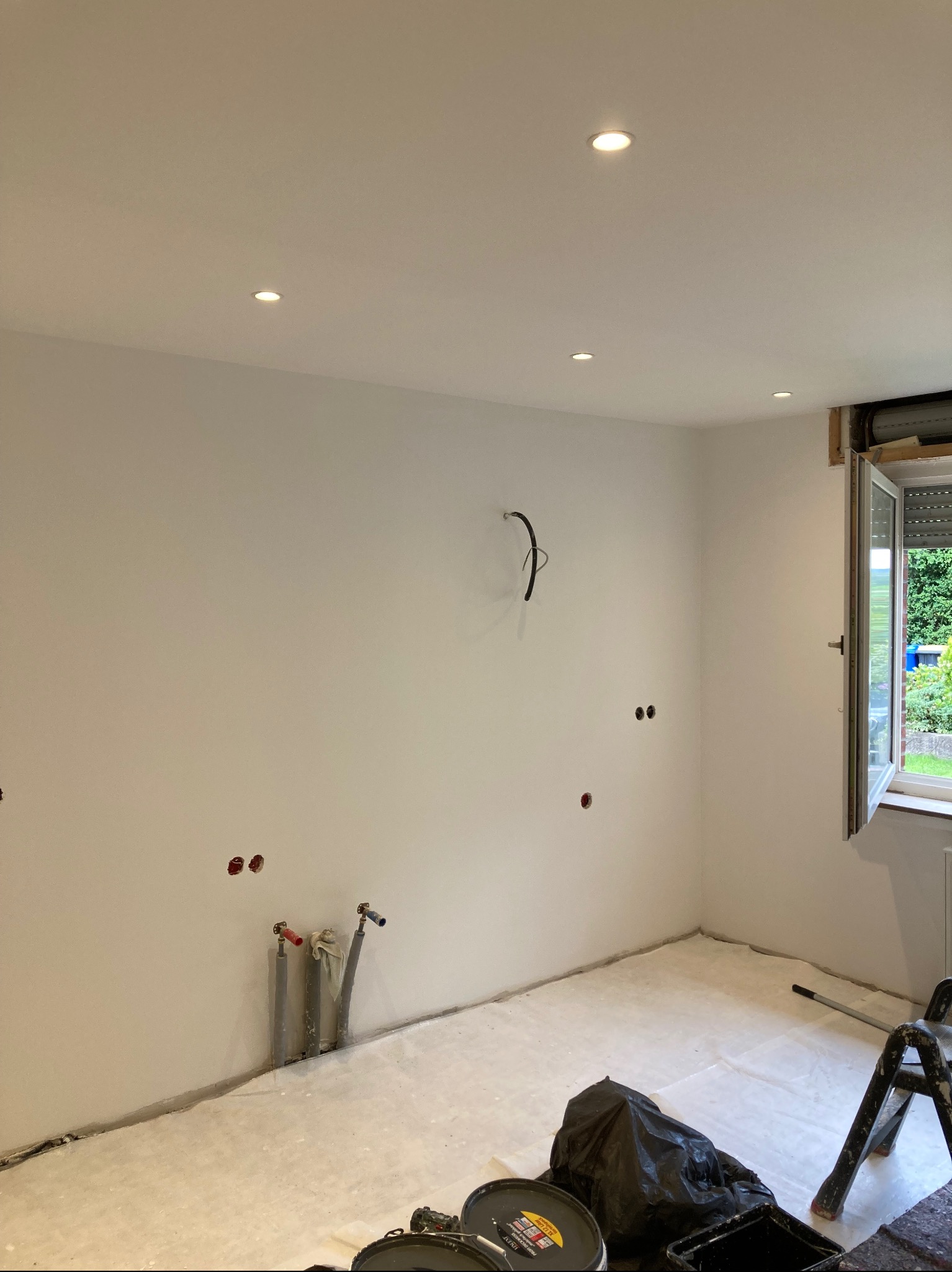 Wnętrze pokoju po świeżym pomalowaniu na biało, z widocznymi instalacjami i przygotowaniami do dalszych prac wykończeniowych. Otwarty oknem z widokiem na zieleń.