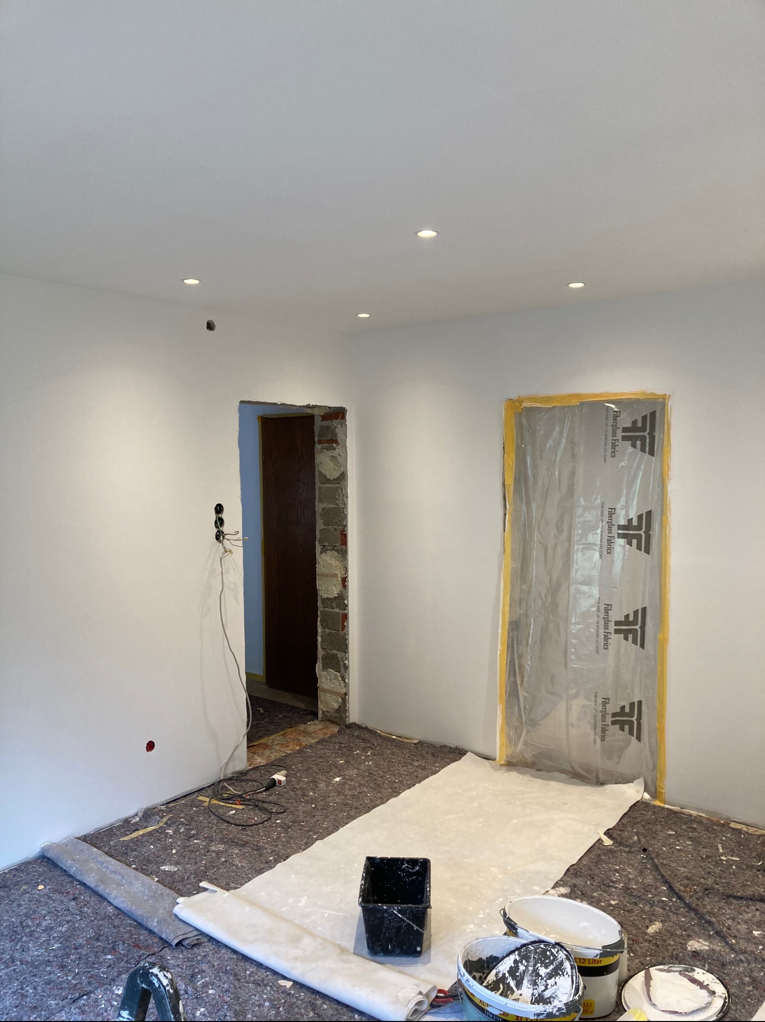 Białe, świeżo pomalowane wnętrze w trakcie remontu, z zabezpieczonymi folią drzwiami i widocznymi narzędziami malarskimi na podłodze. W tle otwór drzwiowy.