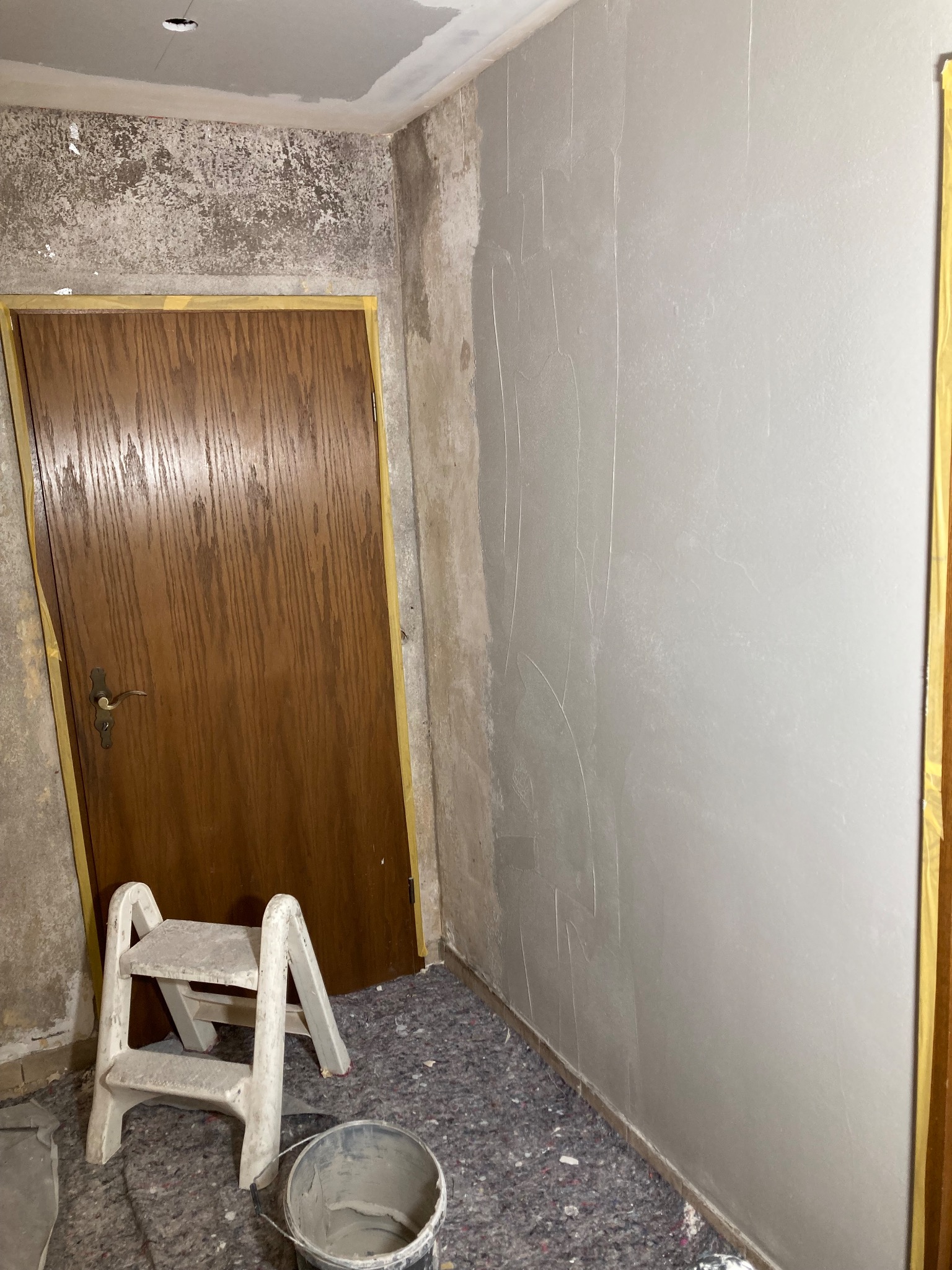 Ściana po szpachlowaniu, z widocznymi śladami po narzędziach. U dołu wiadro i drabinka. Brązowe drzwi z żółtą taśmą zabezpieczającą.