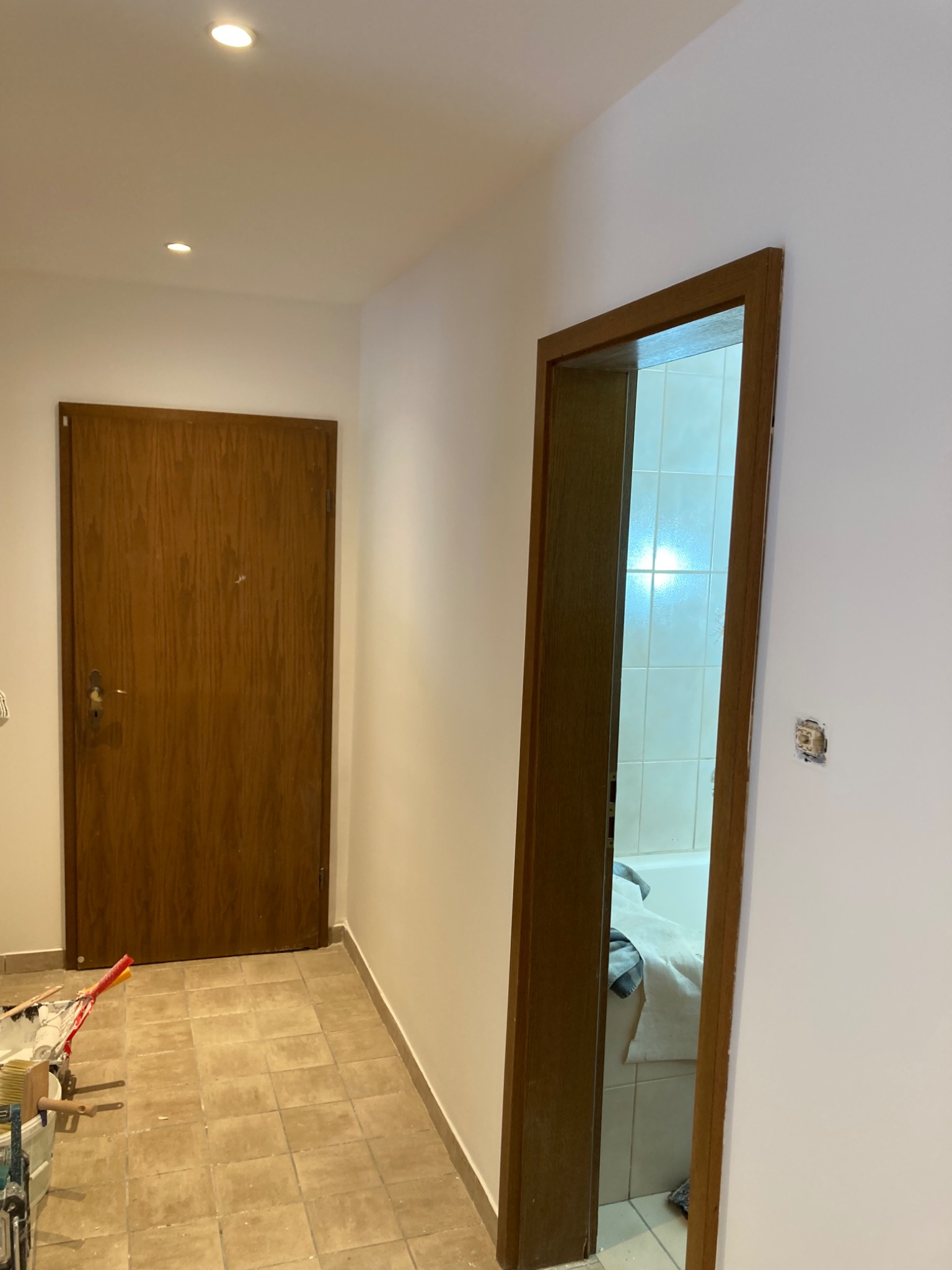 Korytarz po remoncie: świeżo pomalowane ściany na biało, drewniane drzwi wejściowe i do łazienki z widocznymi białymi płytkami. Na podłodze beżowe płytki.