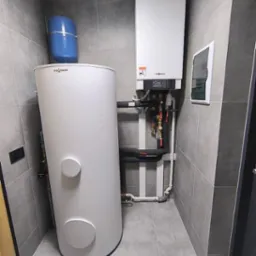 Biały zbiornik akumulacyjny Viessmann obok wiszącego pieca gazowego w pomieszczeniu wyłożonym szarymi płytkami, widoczna instalacja hydrauliczna i skrzynka elektryczna, na podłodze kratka ściekowa.
