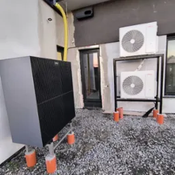 Dwa zewnętrzne moduły klimatyzacji i duża, czarna pompa ciepła Viessmann na zewnątrz budynku, zamontowane na betonowych postumentach, z widoczną rurą spustową i oknami z odbiciem zachodzącego słońca.