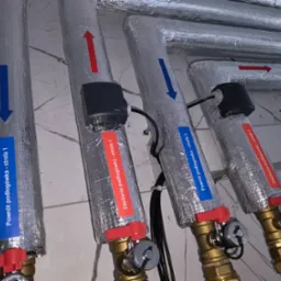Izolowane rury instalacji podłogowej z oznaczeniami stref, zaworami i czujnikami, widoczne strzałki wskazujące kierunek przepływu, etykiety 'powrót' i 'zasilanie'.