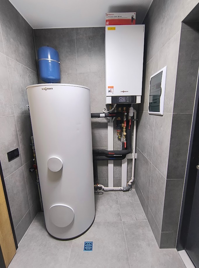 Biały zbiornik akumulacyjny Viessmann obok wiszącego pieca gazowego w pomieszczeniu wyłożonym szarymi płytkami, widoczna instalacja hydrauliczna i skrzynka elektryczna, na podłodze kratka ściekowa.