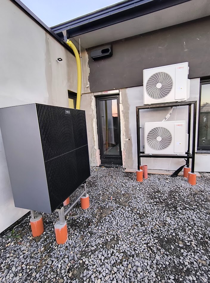 Dwa zewnętrzne moduły klimatyzacji i duża, czarna pompa ciepła Viessmann na zewnątrz budynku, zamontowane na betonowych postumentach, z widoczną rurą spustową i oknami z odbiciem zachodzącego słońca.