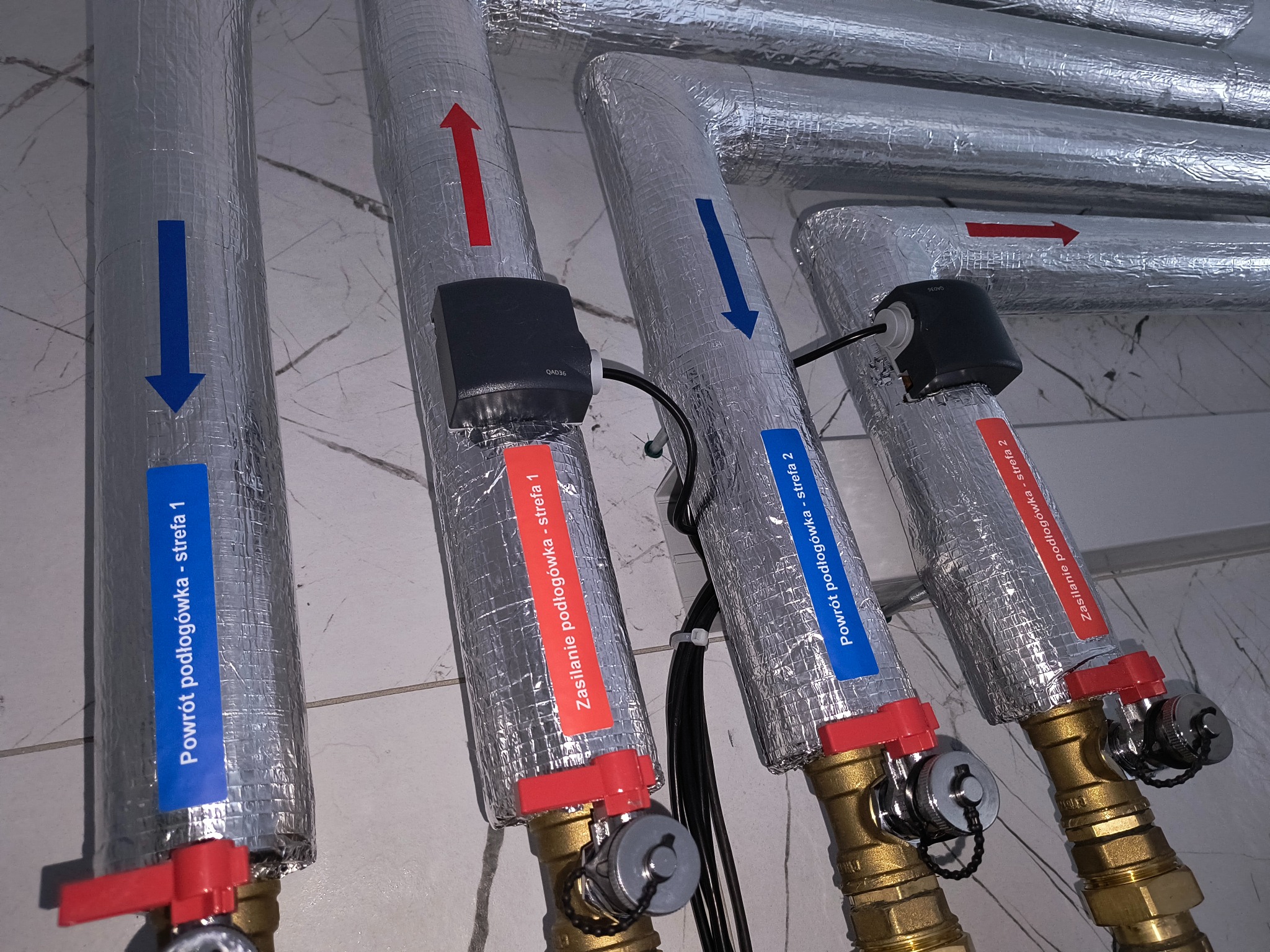Izolowane rury instalacji podłogowej z oznaczeniami stref, zaworami i czujnikami, widoczne strzałki wskazujące kierunek przepływu, etykiety 'powrót' i 'zasilanie'.