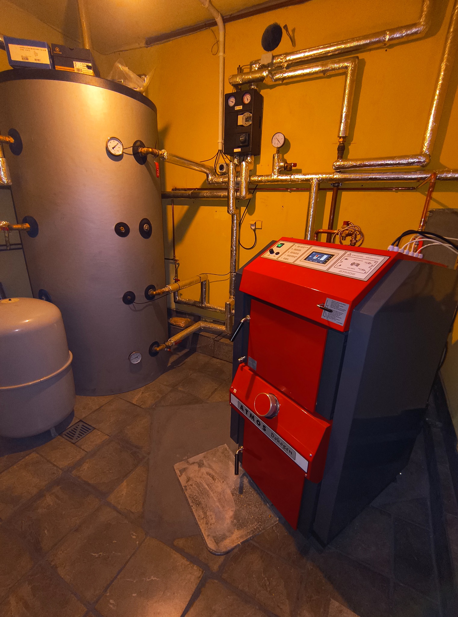Kotłownia z czerwonym piecem Atmos Dokogen, zasobnikiem ciepłej wody i rurami w izolacji na żółtej ścianie.