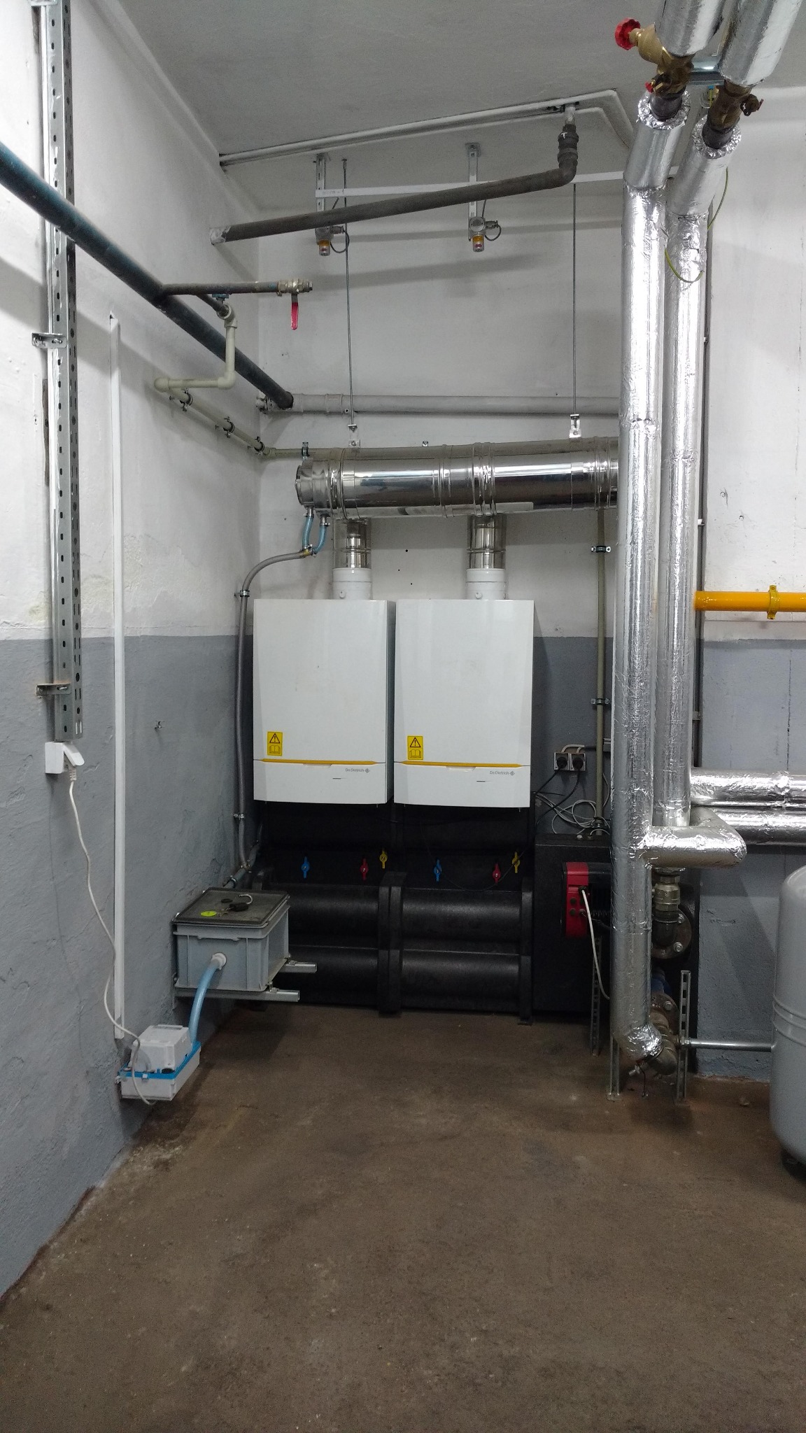 Dwa białe piece grzewcze z żółtymi ostrzeżeniami, podłączone do systemu rur i zbiorników w piwnicy z szarą lamperią.