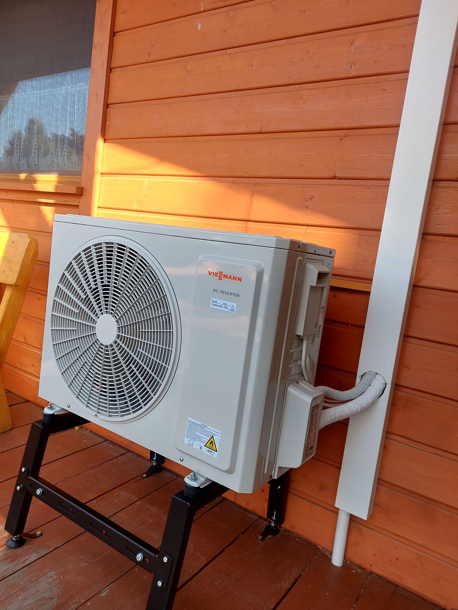 Zewnętrzna jednostka klimatyzacji Viessmann DC Inverter zamontowana na czarnej metalowej podstawie, przy drewnianej ścianie budynku z widoczną białą maskownicą przewodów.
