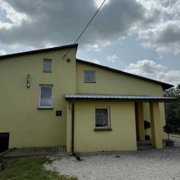 Adam Szmidt - Żółta elewacja domu jednorodzinnego z brązowymi elementami wykończeniowymi, widok z zewnątrz w pochmurny dzień. Widoczne rynny i fragment dachu.