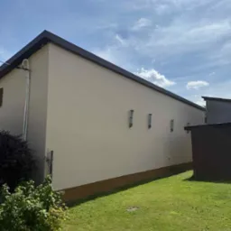 Jasna elewacja budynku z brązowym pasem u dołu, widoczne rury spustowe i fragment dachu. Błękitne niebo z chmurami w tle, trawnik i krzewy na pierwszym planie.