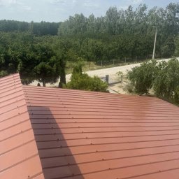 Adam Szmidt - Czerwono-brązowy dach z blachy trapezowej, widok z góry na tle zieleni drzew i błękitnego nieba z chmurami. Nowe pokrycie dachowe, równomierne ułożenie blach.