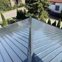 Adam Szmidt - Błyszczący, szary dach z blachy trapezowej, widok z góry, z wyraźnie zaznaczonymi łączeniami. W tle zabudowa jednorodzinna i zieleń.