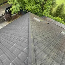 Ciemny, bitumiczny dach skośny z widocznymi zabrudzeniami i mchem, sfotografowany z g&oacute;ry na tle zieleni drzew i zabudowań. Widoczne okna dachowe i komin.