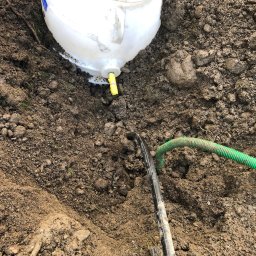 Der-bud inst - Biały zbiornik z żółtym zaworem wystający z wykopu w ziemi, z podłączonymi czarnymi i zielonymi rurami.