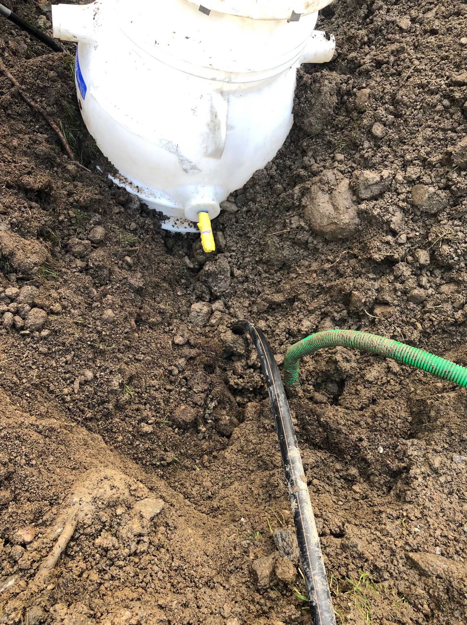 Biały zbiornik z żółtym zaworem wystający z wykopu w ziemi, z podłączonymi czarnymi i zielonymi rurami.