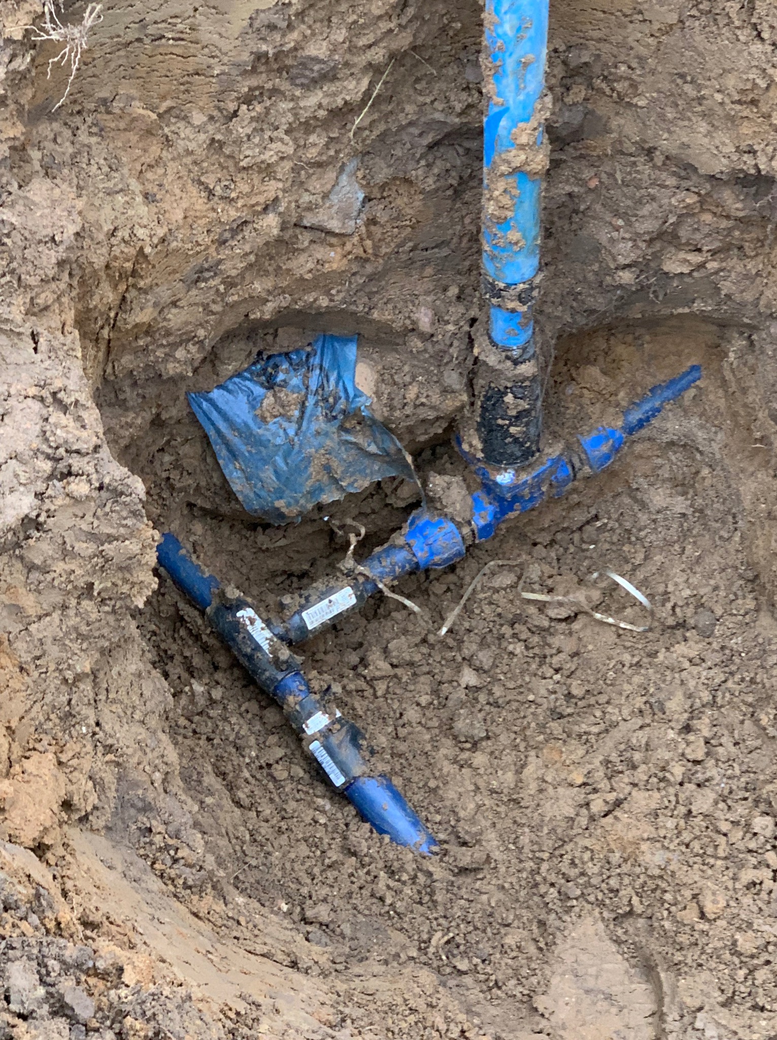Wykop z widocznymi niebieskimi rurami wodociągowymi z łącznikami i elementami montażowymi, częściowo zasypanymi ziemią, oraz fragmentem niebieskiej folii ochronnej.