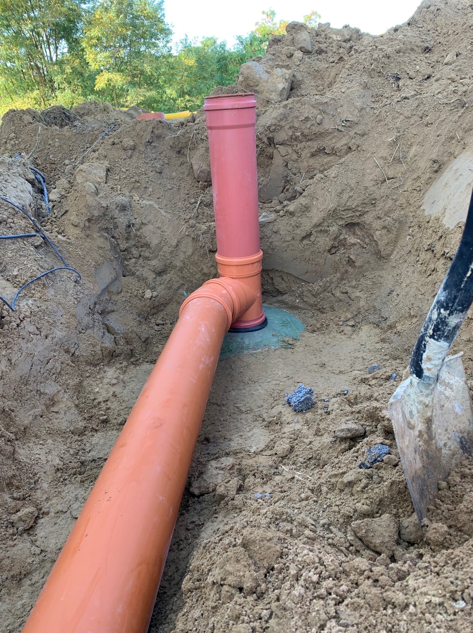 Pomarańczowe rury kanalizacyjne w wykopie, widoczna łopata i kable w ziemi.