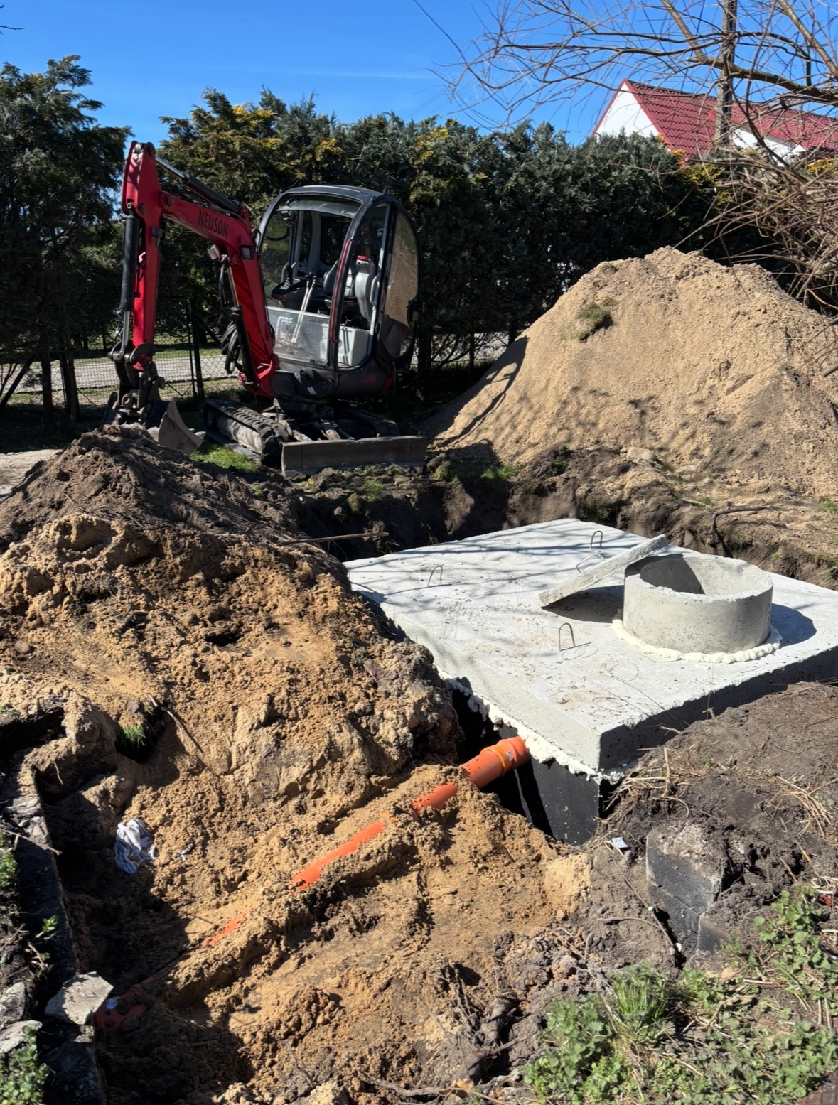 Mini koparka Neuson obok betonowego zbiornika z pomarańczową rurą w wykopie, pryzma ziemi w tle.