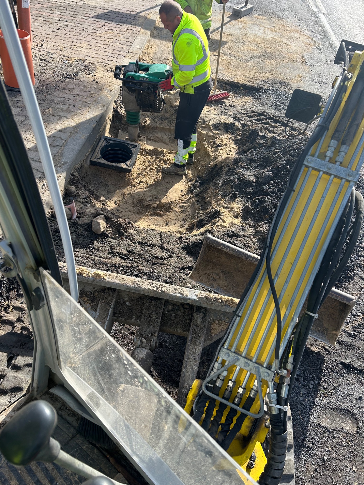 Prace ziemne: ubijanie gruntu zagęszczarką skoczkową przy studzience kanalizacyjnej. Widok z kabiny minikoparki, pracownik w odblaskowej kamizelce.