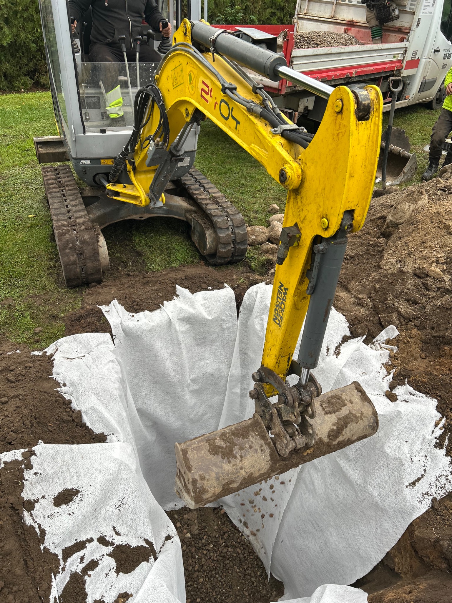 Żółta mini koparka Wacker Neuson w trakcie prac ziemnych, wykop wyłożony białą geowłókniną. W tle widoczny samochód z ładunkiem kruszywa.