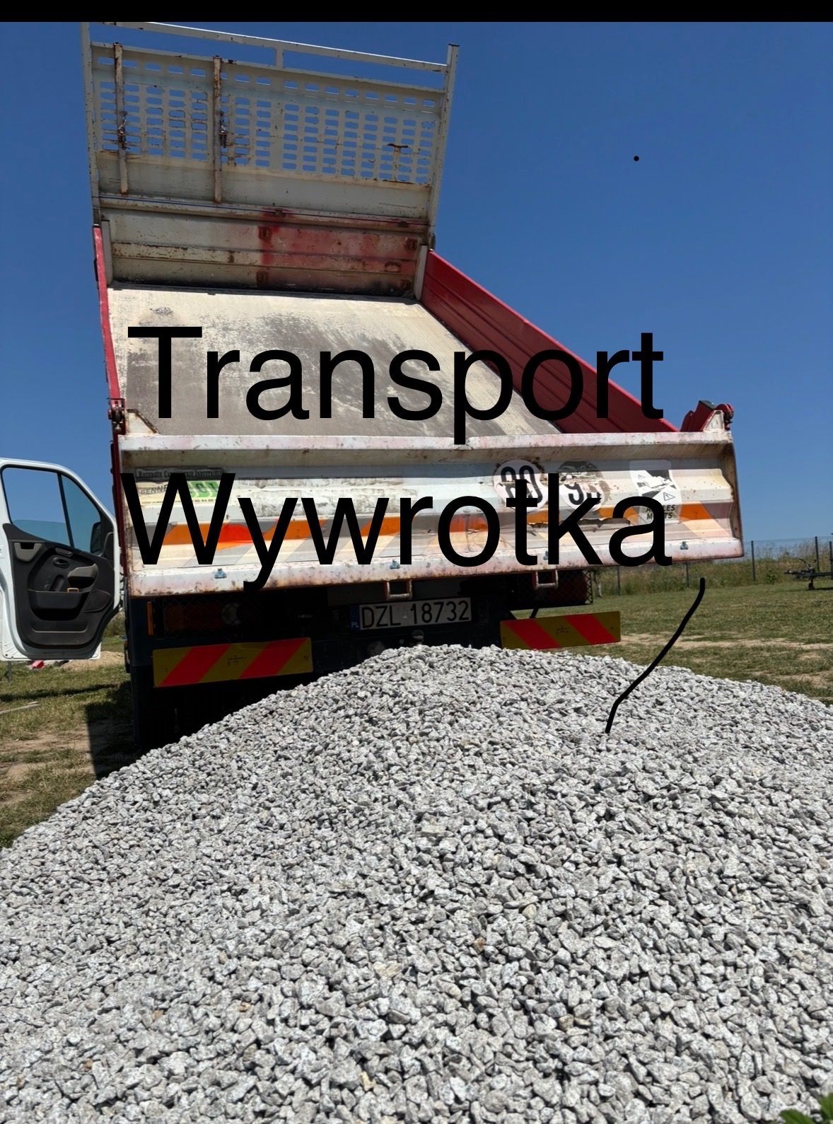 Wywrotka z otwartą skrzynią ładunkową, z której wysypano górę szarego kruszywa na tle błękitnego nieba i zieleni. Widoczny fragment kabiny i tablica rejestracyjna.