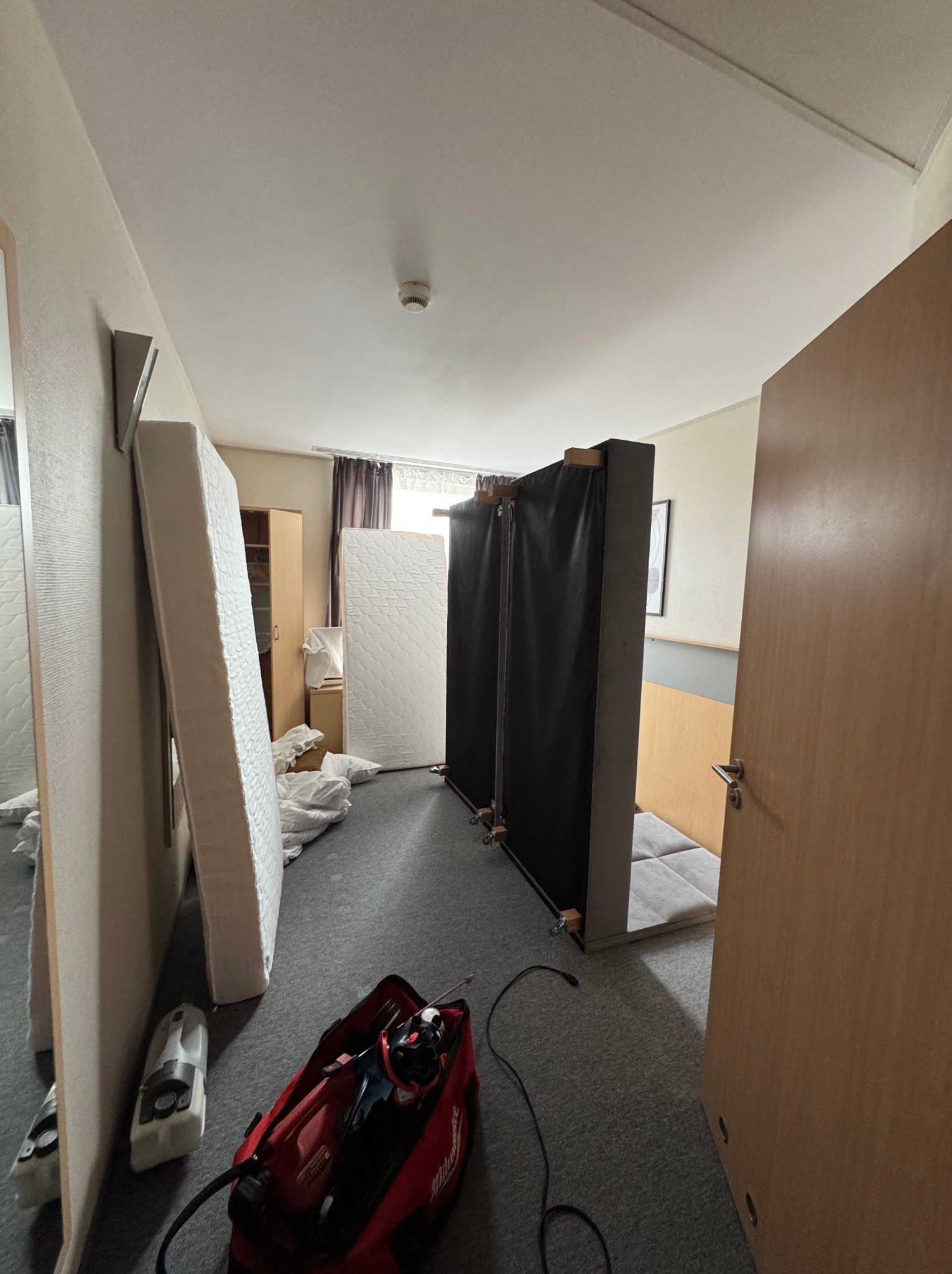 Wnętrze pokoju w trakcie opróżniania: widoczne materace oparte o ściany, rozłożone pościele, sprzęt w torbie i odkurzacz na podłodze.