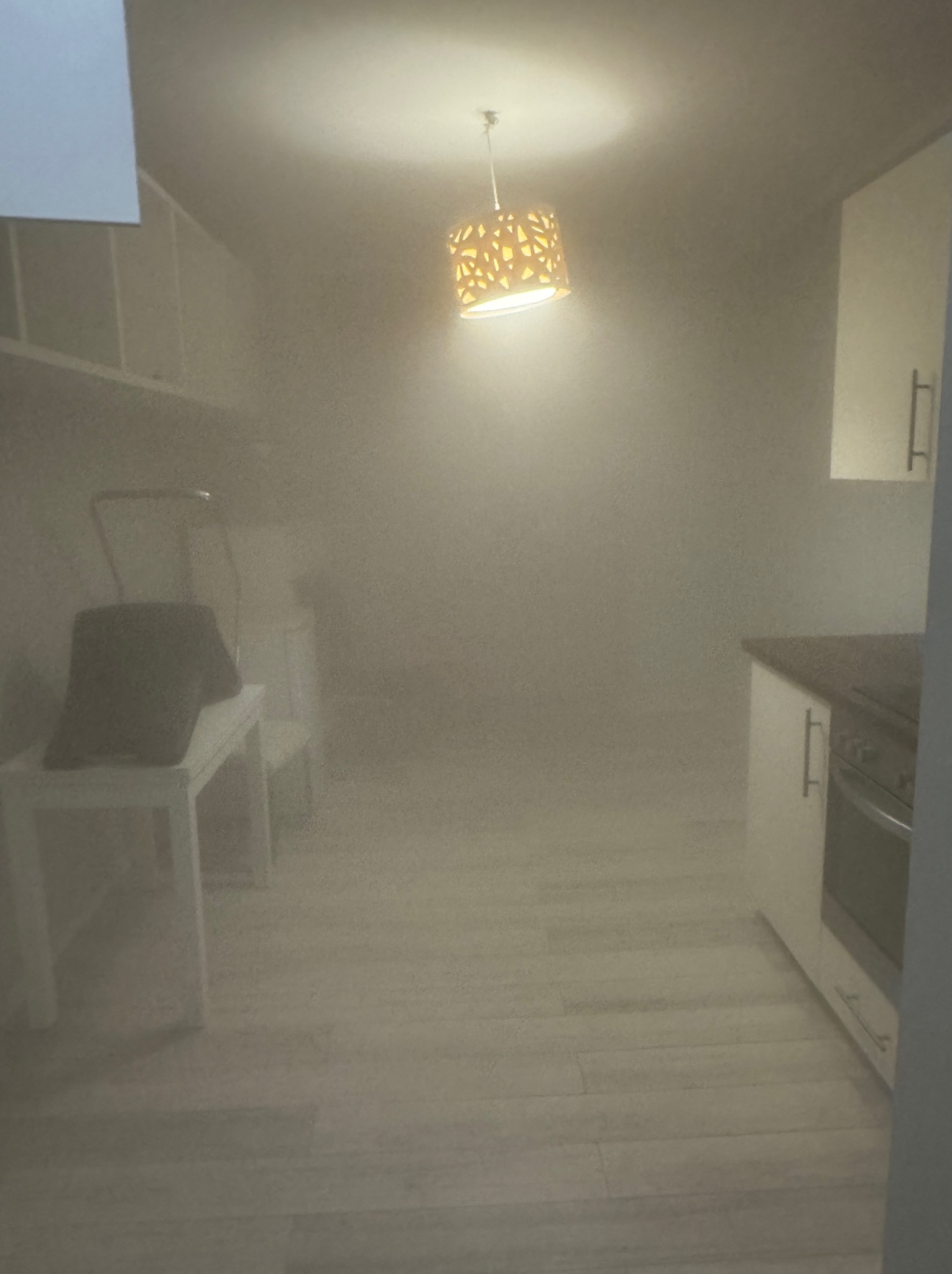 Silne zamglenie w pomieszczeniu z widocznymi meblami kuchennymi, krzesłem i lampą wiszącą, prawdopodobnie po zabiegu czyszczenia lub dezynfekcji.