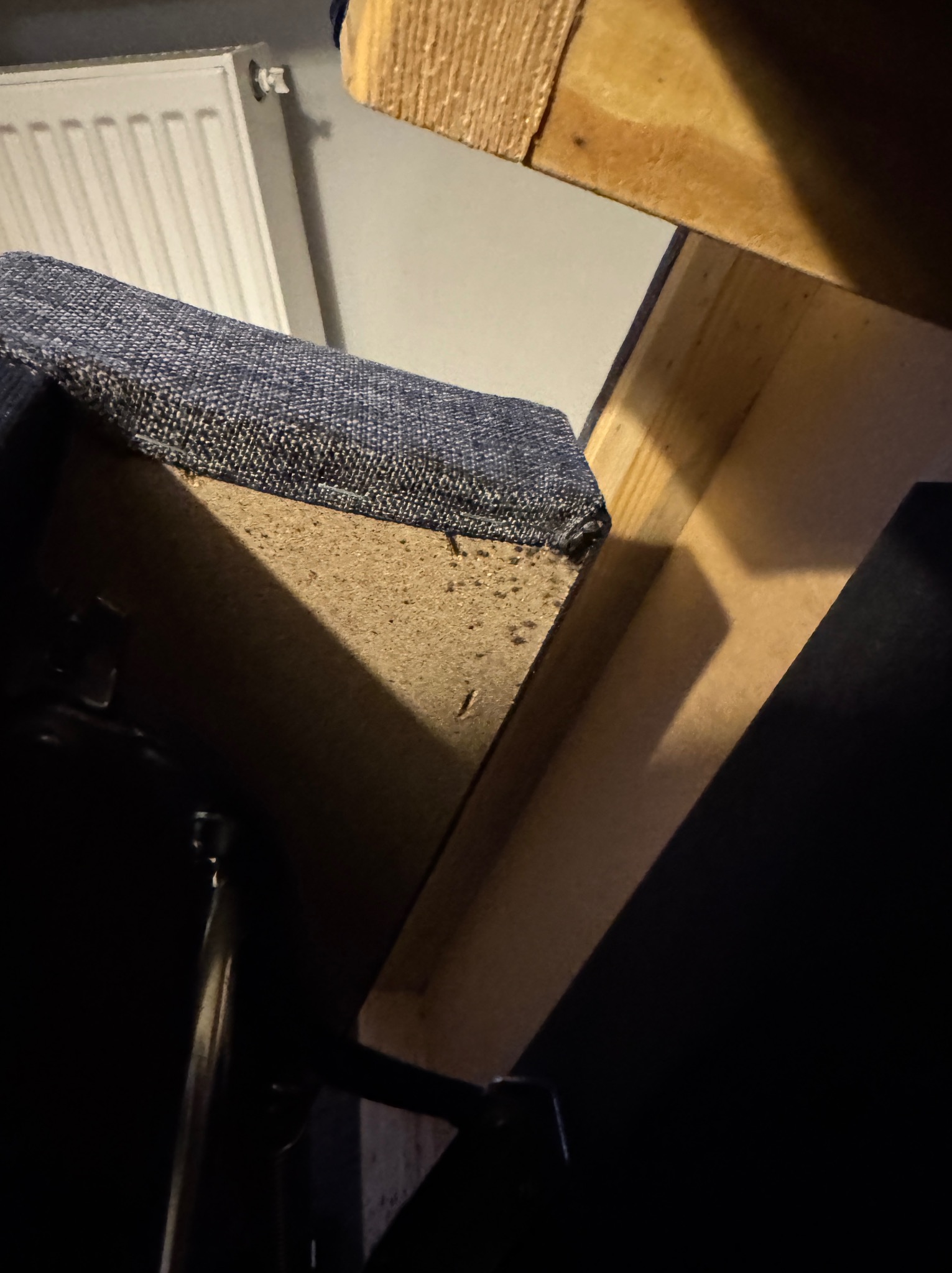 Fragment wnętrza szafki lub schowka z widocznym obiciem siedziska krzesła, drewnianą konstrukcją i fragmentem grzejnika w tle.