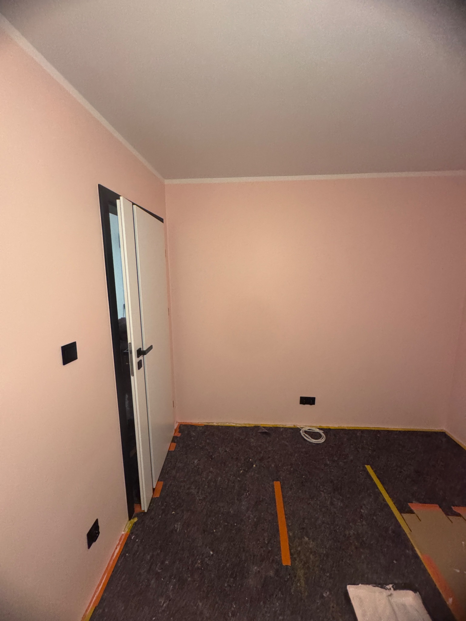 Pokój w trakcie przygotowania do położenia paneli: widoczna podłoga z izolacją, taśmy malarskie przy ścianach, częściowo ułożone panele i białe kable na podłodze.