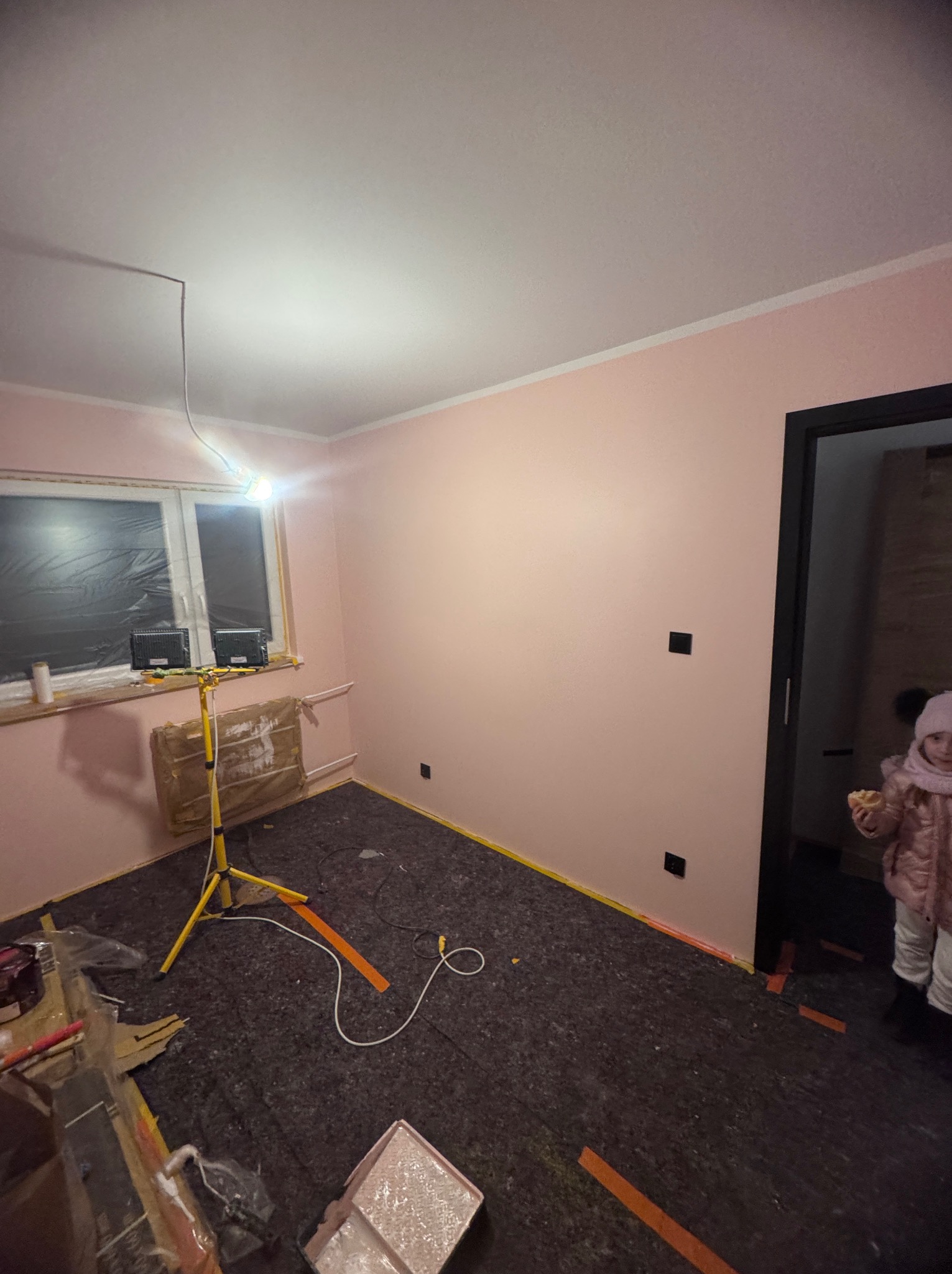 Pokój po malowaniu na różowo, zabezpieczona podłoga, lampa budowlana, widoczne puszki elektryczne, dziecko w drzwiach.