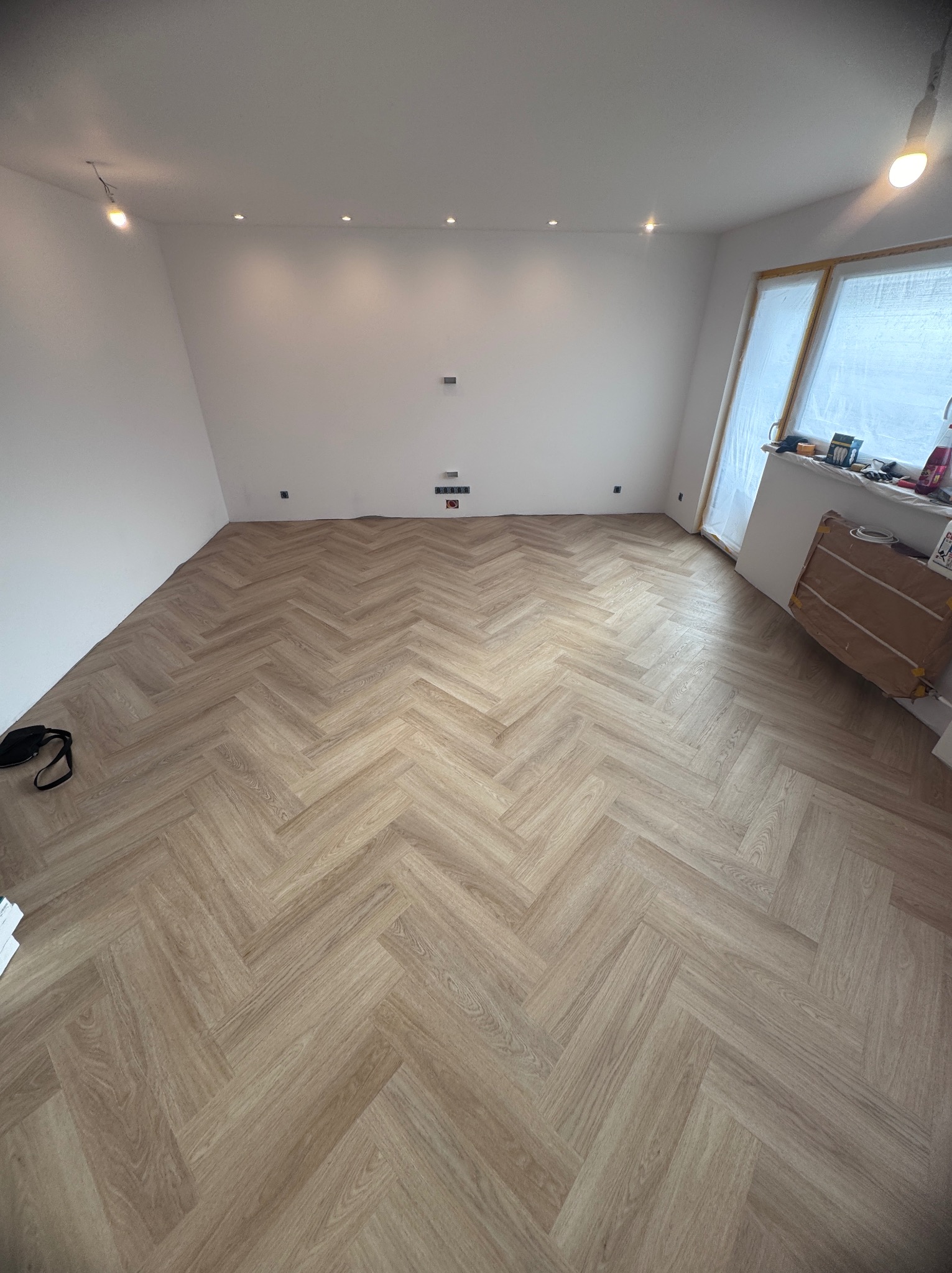 Jasne panele ułożone w jodełkę w pustym, białym pokoju z oknem osłoniętym folią. Widoczne gniazdka elektryczne na ścianie i torba narzędziowa w rogu.