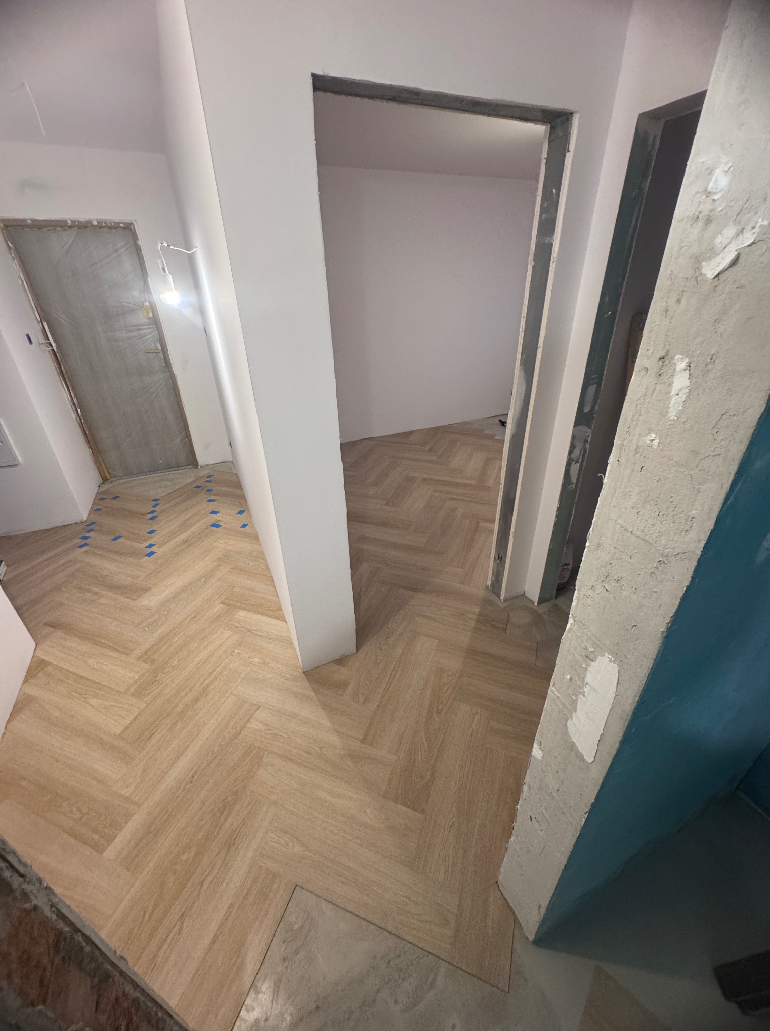 Ułożone panele podłogowe w jodełkę w trakcie prac wykończeniowych, widoczne niezagruntowane futryny drzwiowe i zabezpieczone folią drzwi wejściowe.