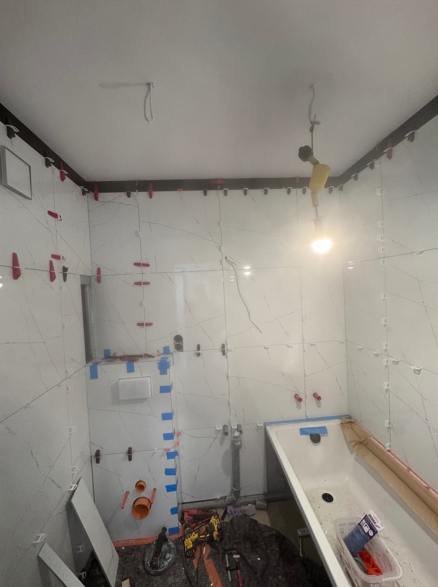 Łazienka w trakcie wykańczania: białe płytki imitujące marmur, system poziomowania płytek, przygotowana instalacja wodno-kanalizacyjna, częściowo zabudowana wanna.