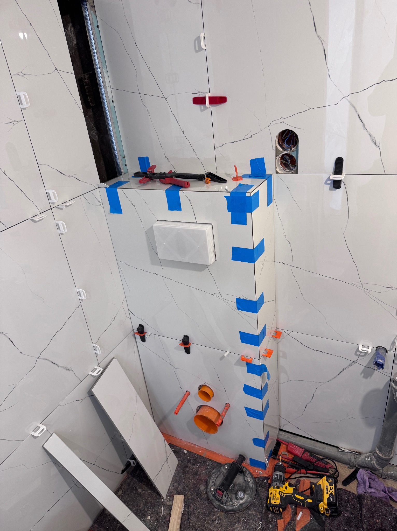 Białe płytki imitujące marmur z czarnymi żyłkami na ścianie łazienki w trakcie montażu, zabezpieczone niebieską taśmą i plastikowymi krzyżykami dystansowymi, widoczne narzędzia glazurnicze i otwory...