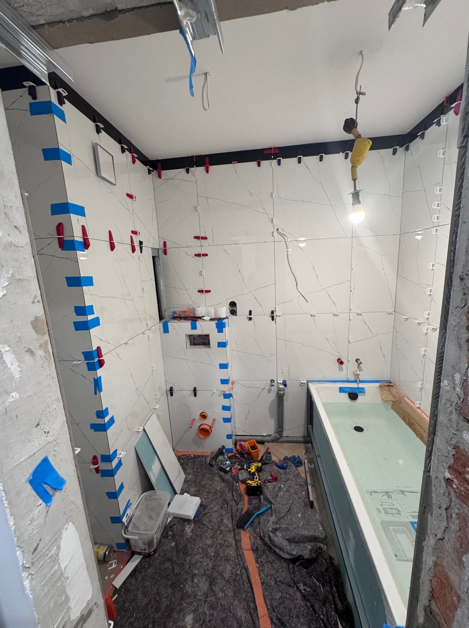 Łazienka w trakcie prac wykończeniowych: białe płytki imitujące marmur z niebieską taśmą i czerwonymi krzyżykami poziomującymi, widoczna wanna wypełniona wodą, narzędzia i materiały budowlane...