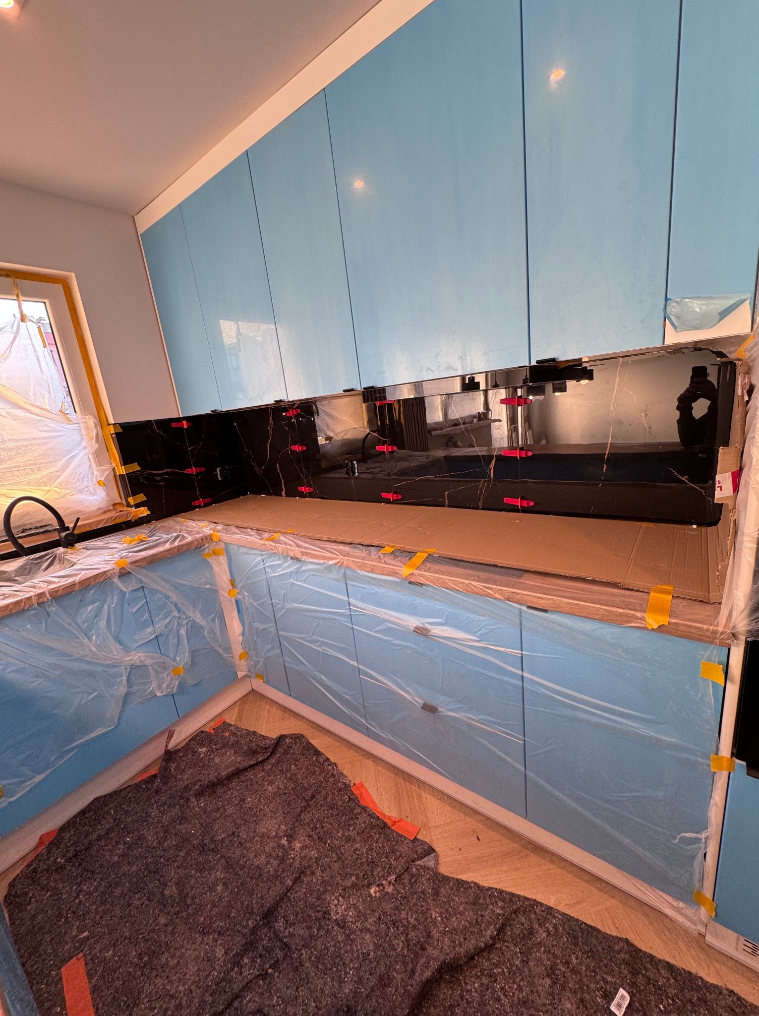 Montaż czarnego panelu ściennego z marmurowym wzorem w kuchni, zabezpieczone folią meble, widoczne czerwone krzyżyki dystansowe i żółta taśma malarska.