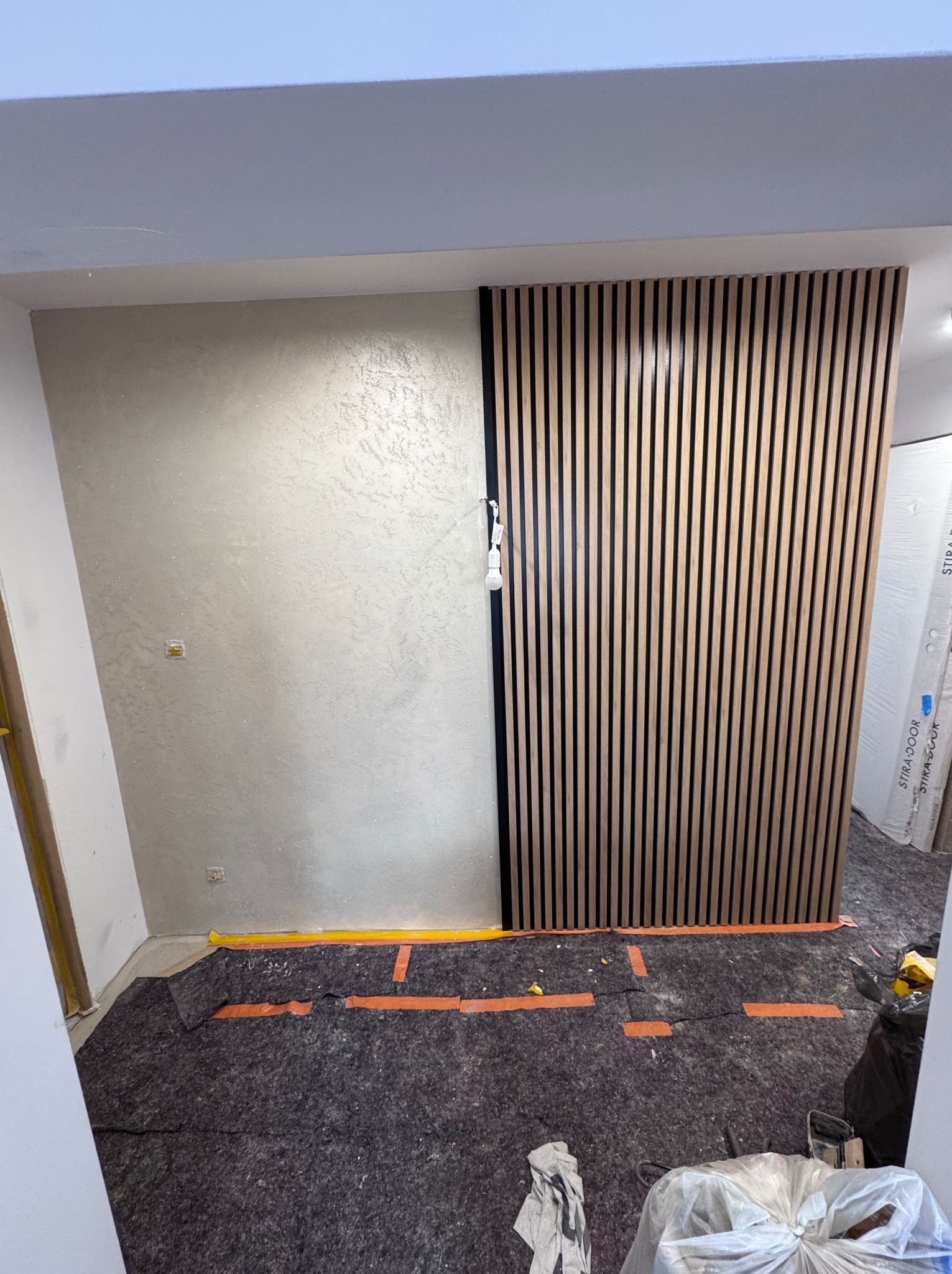 Ściana wykończona dekoracyjnym tynkiem strukturalnym sąsiadująca z drewnianą ścianką działową z pionowych listew, podłoga zabezpieczona materiałem ochronnym podczas prac wykończeniowych.