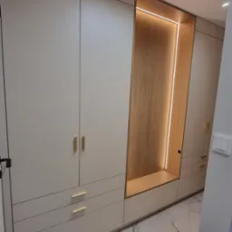 Elegancka szafa wnękowa z beżowymi frontami, złotymi uchwytami i podświetlaną wnęką z drewnianym wykończeniem. Marmurowa podłoga i oświetlenie punktowe.