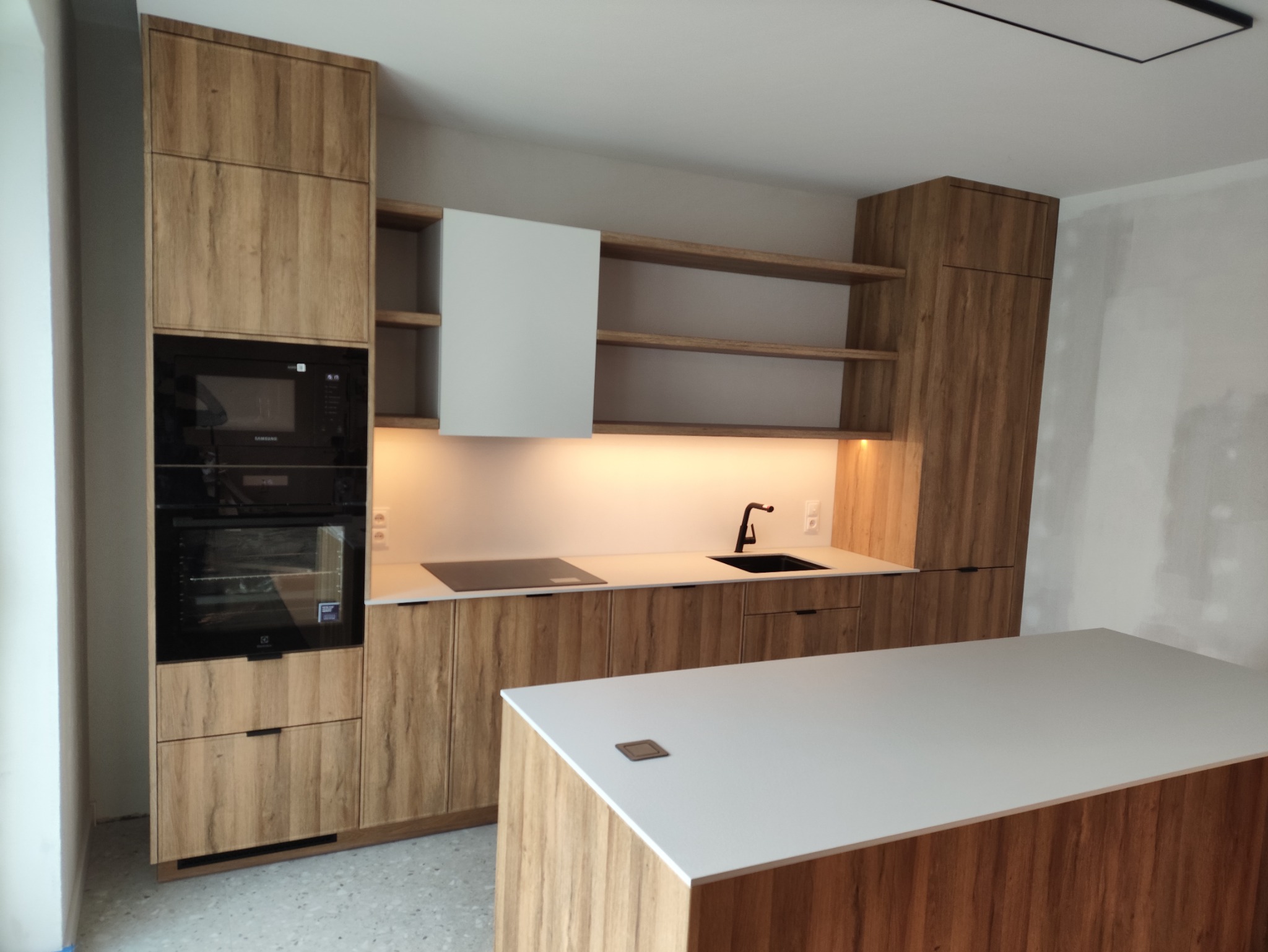 Nowoczesna kuchnia z drewnianymi frontami, białym blatem wyspowym i zabudowanymi sprzętami AGD. Widoczne otwarte półki i oświetlenie podszafkowe.